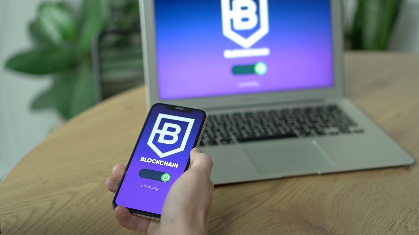 A person holding a smartphone beside a laptop showing an illustrative blockchain screen